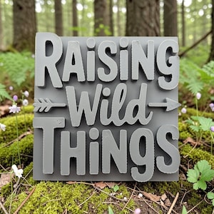 May include: A gray wooden sign with the words "RAISING WILD THINGS" in a layered, 3D effect. An arrow design is between the words "Wild" and "Things". The sign is displayed outdoors, surrounded by greenery and wildflowers, suggesting a nature-themed decor item.