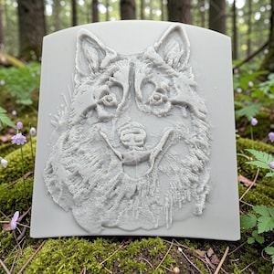 May include: A light gray rectangular plaque featuring a detailed relief of a dog's face. The dog has a fluffy coat, pointed ears, and a smiling expression. The plaque is set against a natural background of green foliage and trees.
