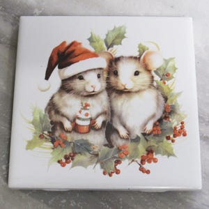 Può includere: Piastrella quadrata in ceramica con due topi. Uno indossa un cappello da Babbo Natale e tiene in mano un cupcake. L'immagine è circondata da agrifoglio e bacche rosse, a tema natalizio.