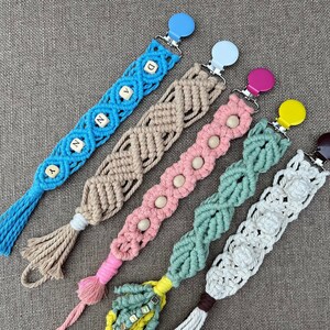 Peut inclure: Cinq attaches-tétines en crochet de couleurs vives avec différents embellissements. Les attaches sont faites de différentes couleurs, notamment le bleu, le rose, le vert et le blanc. Les attaches sont décorées de perles en bois, de perles en lettres et de petits pompons en crochet.