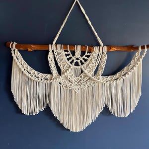 May include: A white macrame wall hanging with a wooden dowel rod and fringe. The hanging features a geometric pattern and is attached to a blue wall.