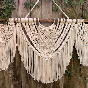 Puede incluir: Un tapiz de macramé blanco con cuentas de madera y flecos. El tapiz está formado por tres piezas separadas, cada una con un patrón diferente. El tapiz se muestra sobre un fondo de madera.