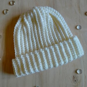 Puede incluir: Un gorro de punto blanco con textura de canal&eacute;. El gorro est&aacute; sobre una superficie de madera clara con peque&ntilde;as cuentas de vidrio esparcidas a su alrededor.