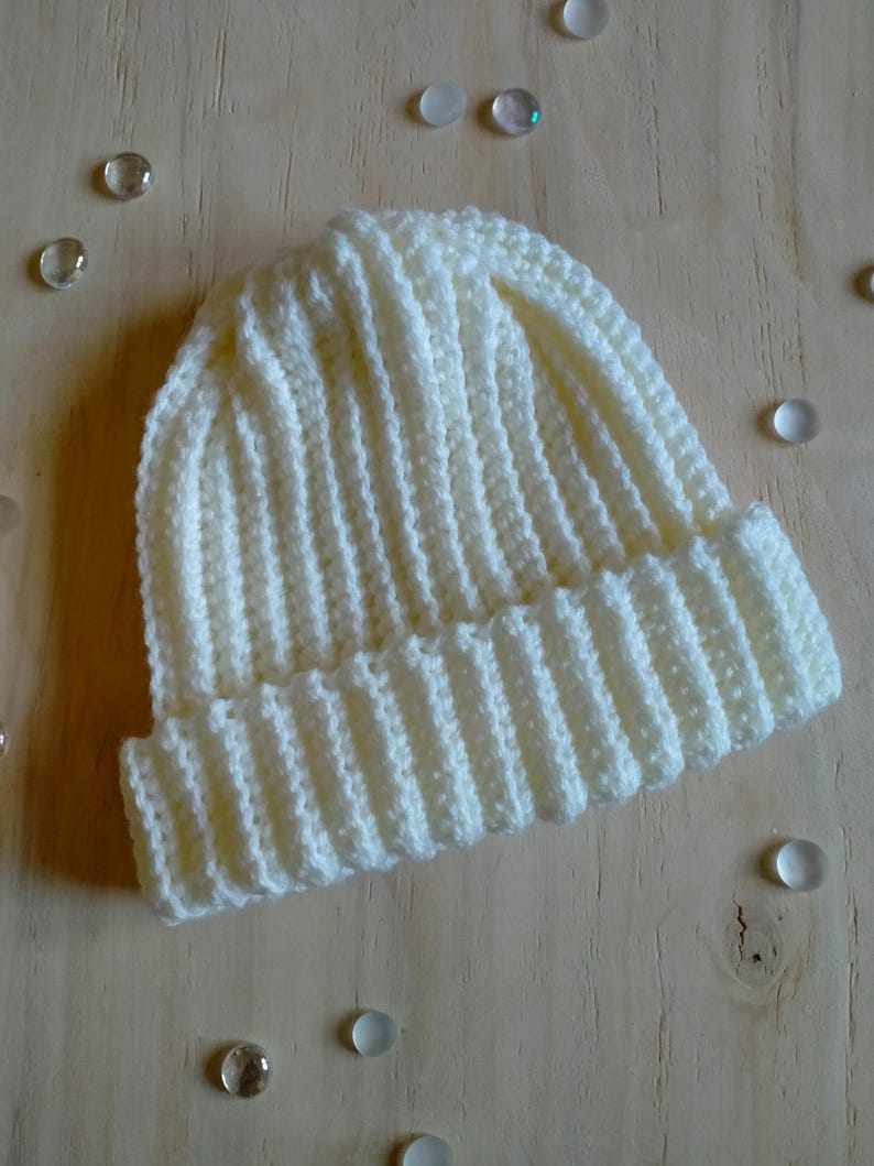 Puede incluir: Un gorro de punto blanco con textura de canal&eacute;. El gorro est&aacute; sobre una superficie de madera clara con peque&ntilde;as cuentas de vidrio esparcidas a su alrededor.