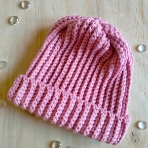 Puede incluir: Un gorro de punto de color rosa sobre un fondo de madera clara con peque&ntilde;as piedras de vidrio esparcidas a su alrededor.