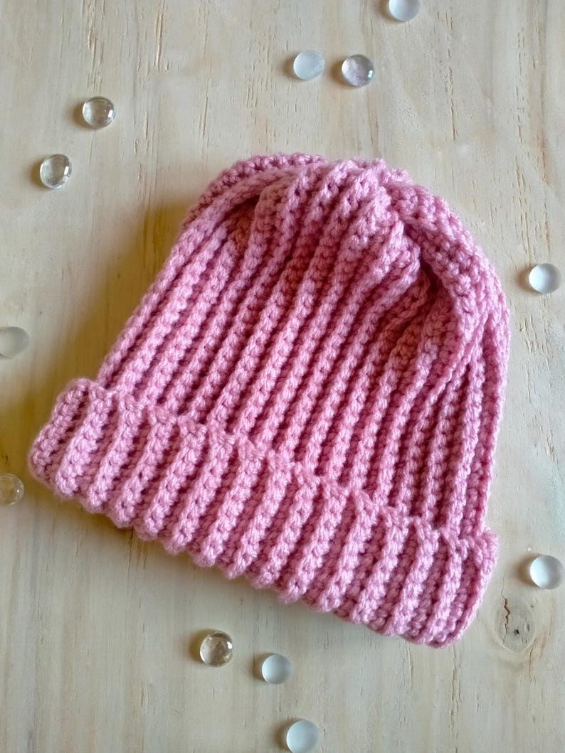 Puede incluir: Un gorro de punto de color rosa sobre un fondo de madera clara con peque&ntilde;as piedras de vidrio esparcidas a su alrededor.