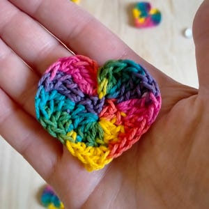 Può includere: Un cuore all'uncinetto arcobaleno con un motivo a spirale. Il cuore è realizzato con filati di diversi colori, tra cui viola, rosa, verde, giallo e arancione.