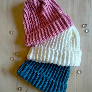 Puede incluir: Tres gorros de punto de ganchillo apilados uno encima del otro. El gorro de arriba es rosa, el del medio es blanco y el de abajo es azul verdoso.
