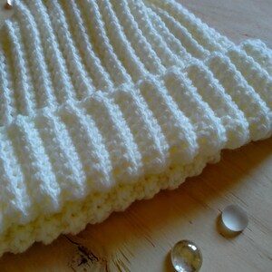 Puede incluir: Un gorro de punto blanco con textura de canal&eacute;. El gorro est&aacute; sobre una superficie de madera marr&oacute;n claro.