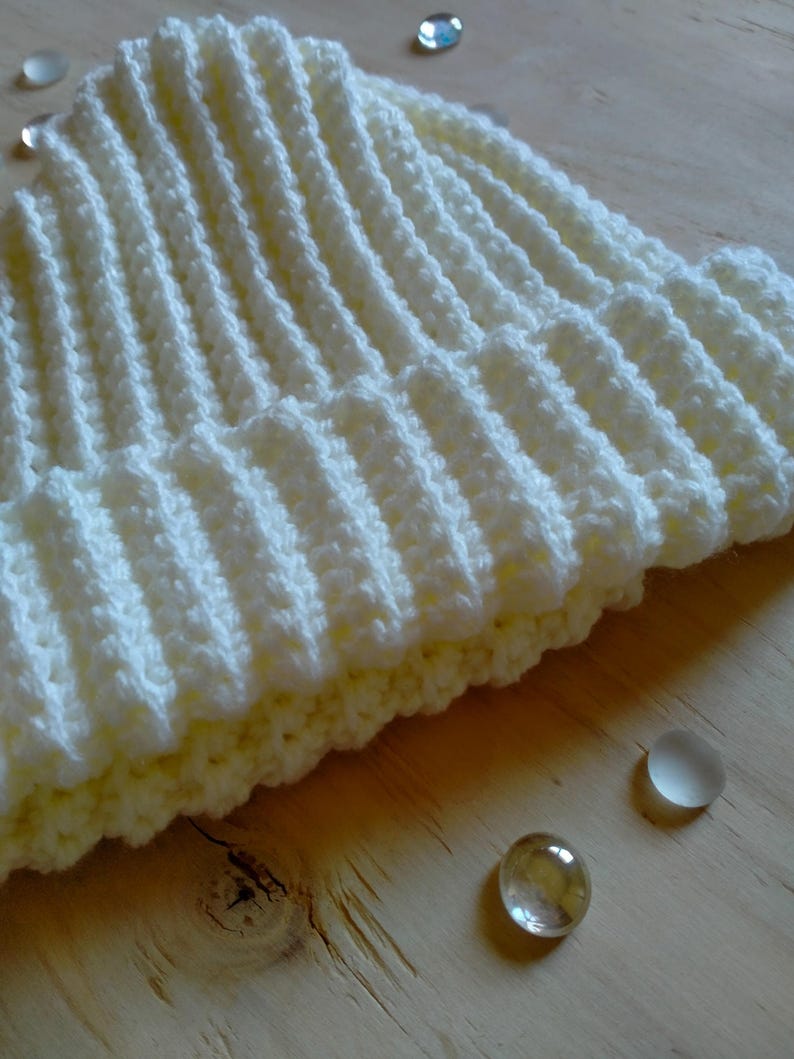 Puede incluir: Un gorro de punto blanco con textura de canal&eacute;. El gorro est&aacute; sobre una superficie de madera marr&oacute;n claro.