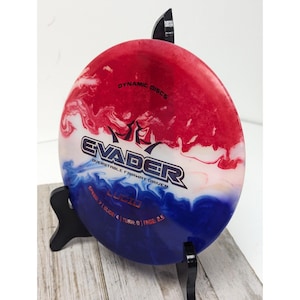 May include: A red, white, and blue Dynamic Discs Evader overstable fairway driver. The disc has the word "Evader" in large black letters, with the word "Lucid" below. The disc is on a black stand.