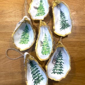 May include: Six oyster shell Christmas tree ornaments. Each shell has a gold-painted rim and a painted green Christmas tree design. They have a white interior and a twine loop for hanging. The shells are on a wooden surface.