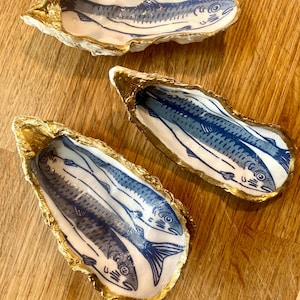 May include: Three decorative oyster shells featuring hand-painted blue fish illustrations on the interior. Each shell has a scalloped, irregular edge finished with a metallic gold trim. The shells are white with detailed blue ink-style fish motifs.