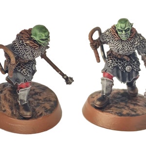 Könnte beinhalten: Zwei grünhäutige Fantasy-Miniaturen von Orks, die Kettenhemden tragen und Waffen halten. Die Orks stehen auf braunen Sockeln.
