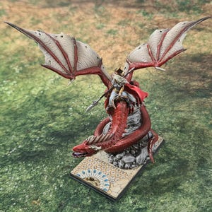 Könnte beinhalten: Eine Miniaturfigur einer Frau, die auf einem roten Drachen mit weißen Flügeln reitet. Der Drache sitzt auf einem Steinsockel mit einem dekorativen Muster. Die Frau trägt ein weißes und goldenes Outfit und hält eine Lanze.