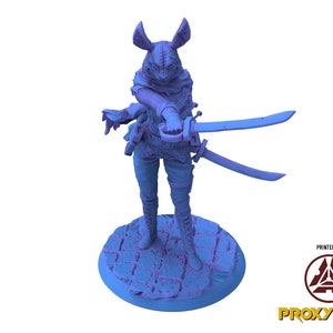 Könnte beinhalten: Eine blaue 3D-gedruckte Miniatur einer weiblichen Figur mit Hasenohren, die einen Kapuzenumhang trägt und zwei Schwerter hält. Die Figur steht auf einem runden Sockel mit einer strukturierten Oberfläche.