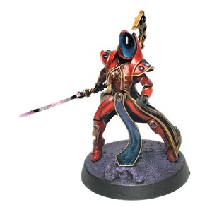 May include: A red and gold painted miniature figure of a warrior holding a sword. The figure is wearing a blue cape and has a blue face mask. The figure is standing on a black base.