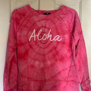 Puede incluir: Una sudadera rosa tie-dye con la palabra "Aloha" bordada en blanco en la parte delantera.