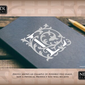 May include: A close-up of a black notebook with a white letter L surrounded by a decorative floral design. The notebook is on a wooden surface with a pencil lying next to it.