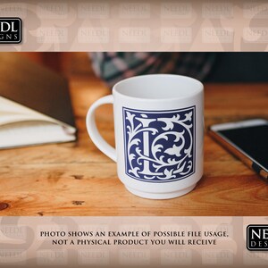 May include: White ceramic mug with a blue and white floral design featuring the letter "I" in the center. The mug is on a wooden table with a notebook and a cell phone.