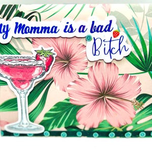 Könnte beinhalten: Eine Grußkarte mit tropischem Thema, mit rosa Hibiskusblüten und grünen Blättern. Die Karte enthält einen Aufkleber einer rosa Margarita mit Erdbeere und dem Text "My Momma is a bad Bitch" in Blau.
