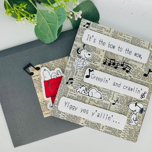 May include: A square greeting card featuring a vintage newspaper print background with Snoopy illustrations, black musical note cutouts, and white text labels reading It's the bow to the wow, Creepin' and crawlin', and Yiggy yes y'allin'.