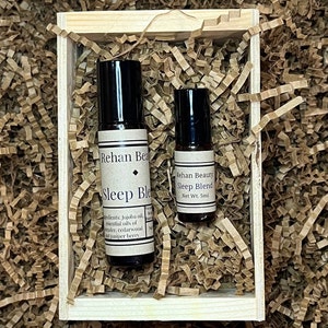 May include: Two small glass bottles with black roll-on tops in a wooden box filled with brown packing paper. The bottles contain essential oil blends for sleep. The labels read "Rehan Beauty Sleep Blend" and "Sleep Blend".