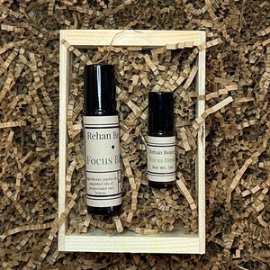 May include: Two small glass bottles with roller balls, both labeled "Rehan Beauty Focus Blend". The larger bottle has additional text listing ingredients. The bottles are in a wooden box with brown paper packing material.