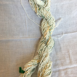 May include: A skein of white yarn with green and yellow flecks. The yarn is twisted and looped, and the ends are tied with green yarn.