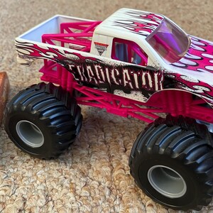 May include: A white and pink monster truck toy with the word "Eradicator" on the side. The truck has large black tires and a pink roll cage. The truck is on a carpeted surface.