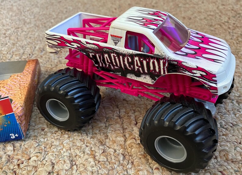 May include: A white and pink monster truck toy with the word "Eradicator" on the side. The truck has large black tires and a pink roll cage. The truck is on a carpeted surface.