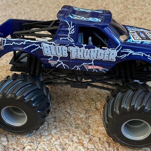 May include: A blue monster truck toy with the words "Blue Thunder" and lightning bolt graphics. The toy truck has large black tyres and a black chassis. The words "Built Tough" are also visible on the side of the truck.