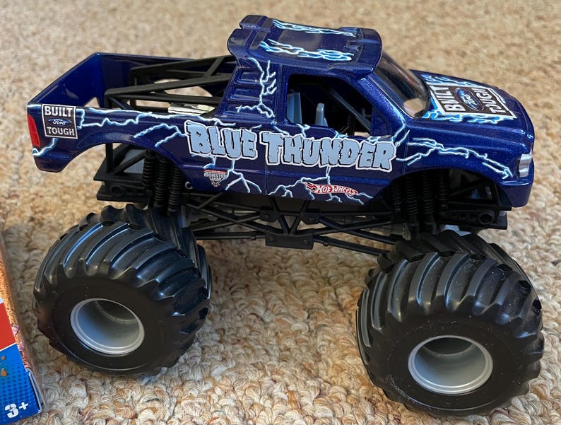 May include: A blue monster truck toy with the words "Blue Thunder" and lightning bolt graphics. The toy truck has large black tyres and a black chassis. The words "Built Tough" are also visible on the side of the truck.