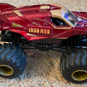 May include: A red and gold Iron Man monster truck toy with large black tires and gold rims. The truck features the Iron Man logo and Stark Industries branding. The toy is designed for off-road play.