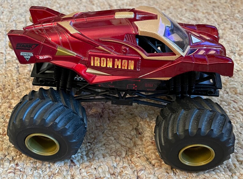 May include: A red and gold Iron Man monster truck toy with large black tires and gold rims. The truck features the Iron Man logo and Stark Industries branding. The toy is designed for off-road play.