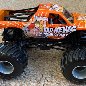 May include: Orange and black toy monster truck with large tires. The truck features the text "BAD NEWS TRAVELS FAST" and a cartoon character graphic. The truck has a black roll cage and is a Hot Wheels product.