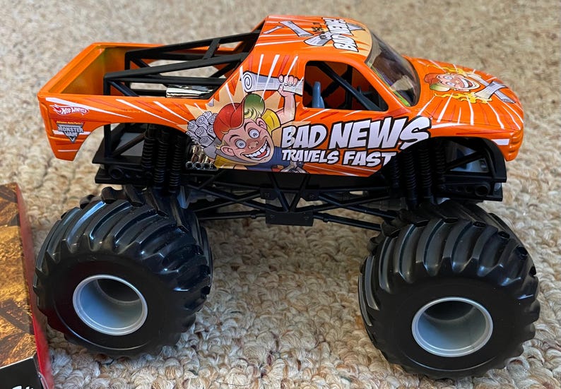 May include: Orange and black toy monster truck with large tires. The truck features the text "BAD NEWS TRAVELS FAST" and a cartoon character graphic. The truck has a black roll cage and is a Hot Wheels product.