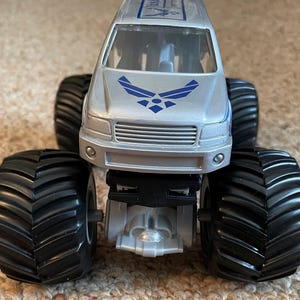 Pu&ograve; includere: Giocattolo di monster truck argentato con grandi pneumatici neri. La parte anteriore del camion presenta il logo dell'US Air Force in blu. Il camion ha una griglia argentata e una striscia blu sulla parte superiore.