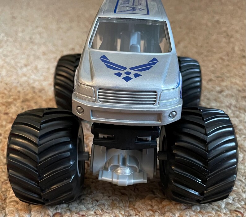 Pu&ograve; includere: Giocattolo di monster truck argentato con grandi pneumatici neri. La parte anteriore del camion presenta il logo dell'US Air Force in blu. Il camion ha una griglia argentata e una striscia blu sulla parte superiore.