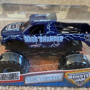 May include: A die-cast metal body Blue Thunder monster truck toy. The dark blue truck features white lightning bolt graphics and the words "Blue Thunder" and "Monster Jam". The toy is packaged in a clear plastic container.