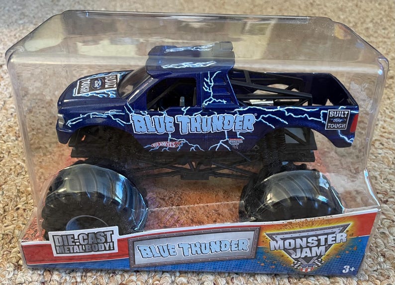 May include: A die-cast metal body Blue Thunder monster truck toy. The dark blue truck features white lightning bolt graphics and the words "Blue Thunder" and "Monster Jam". The toy is packaged in a clear plastic container.