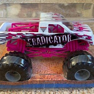 May include: A toy monster truck in a clear plastic case. The truck is white with pink and black flame designs and the word "ERADICATOR" on the side. It has large black tires and pink suspension.