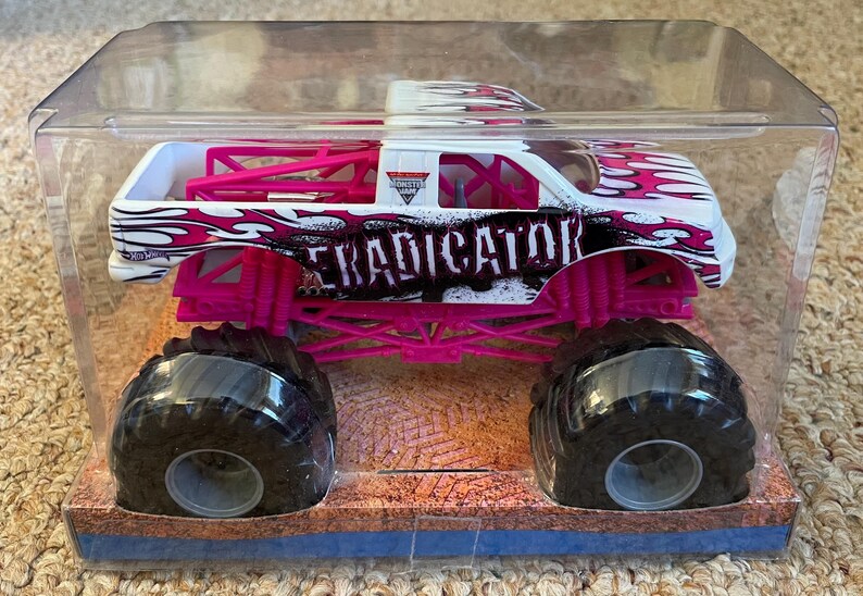 May include: A toy monster truck in a clear plastic case. The truck is white with pink and black flame designs and the word "ERADICATOR" on the side. It has large black tires and pink suspension.