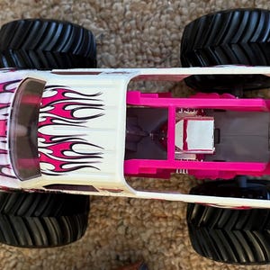 May include: A white and pink monster truck toy with black tires. The truck features pink flame graphics on the side and a pink interior. The large tires are designed for off-road driving.