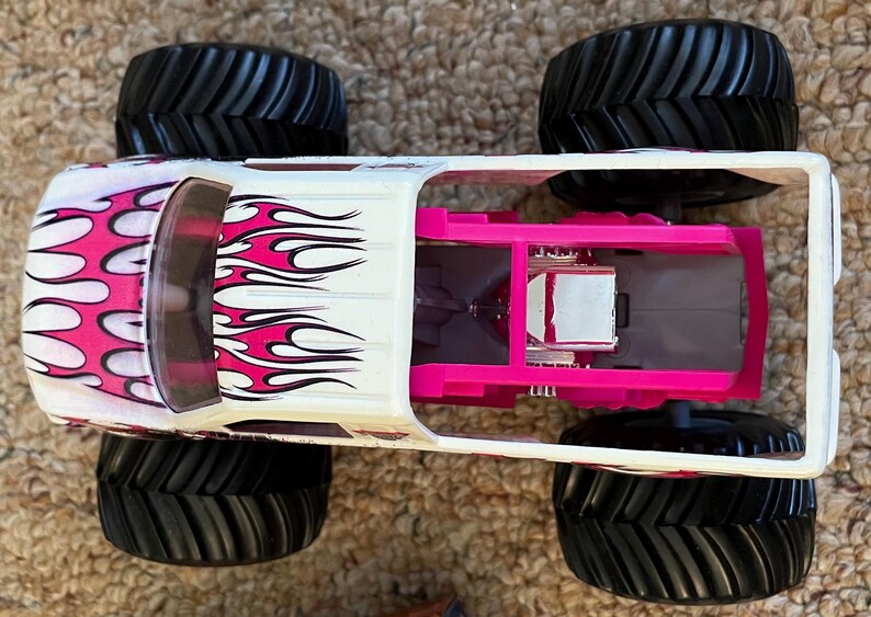 May include: A white and pink monster truck toy with black tires. The truck features pink flame graphics on the side and a pink interior. The large tires are designed for off-road driving.