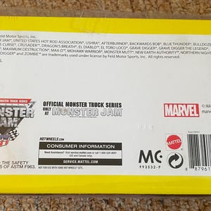 May include: A yellow and white Monster Jam toy package. The package features the Monster Jam logo, the Marvel logo, and the names of various monster trucks. The package also includes consumer information and a barcode.