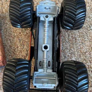 May include: A black and orange monster truck toy with large, textured black tires. The underside of the truck is silver and black, with visible mechanical details. The toy is designed for off-road play.