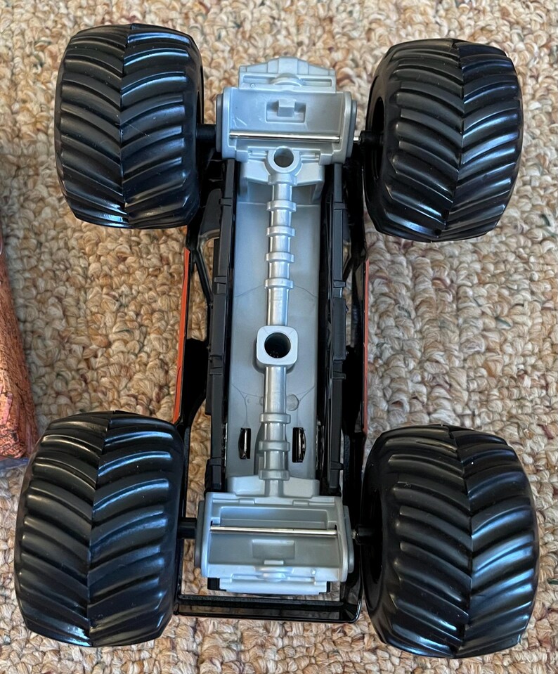May include: A black and orange monster truck toy with large, textured black tires. The underside of the truck is silver and black, with visible mechanical details. The toy is designed for off-road play.