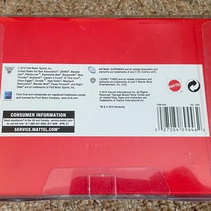 May include: A red box with clear plastic wrapping. The box contains text and logos for Mattel, Ford, and DC Comics. The text includes consumer information and a barcode.