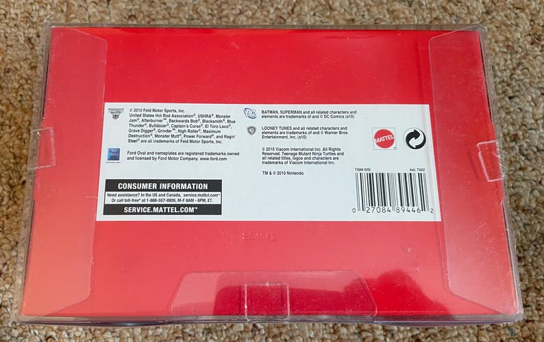 May include: A red box with clear plastic wrapping. The box contains text and logos for Mattel, Ford, and DC Comics. The text includes consumer information and a barcode.